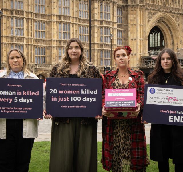 Four women stand outside the houses of parliament with signs and a box calling for an end to violence against women and girls