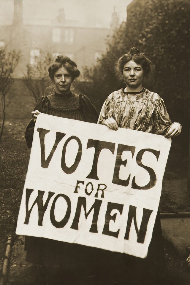 Historic sepia photograph of Annie Kenney and Christabel Pankhurst stood outside holding a giant sign which reads 'VOTES FOR WOMEN'