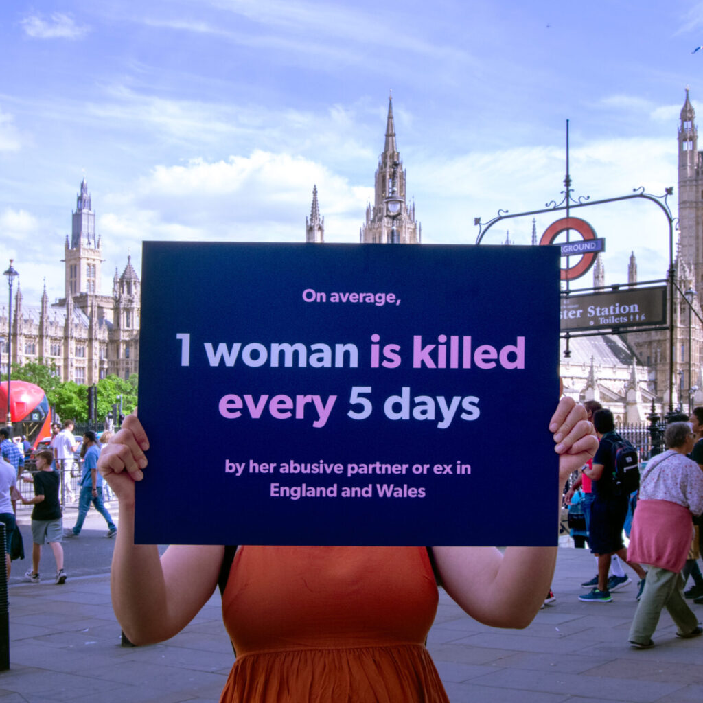 Woman stands outside parliament, holding a placard that covers her face and reads' On average, 1 woman is killed every 5 days by her abusive partner or ex in England and Wales.
