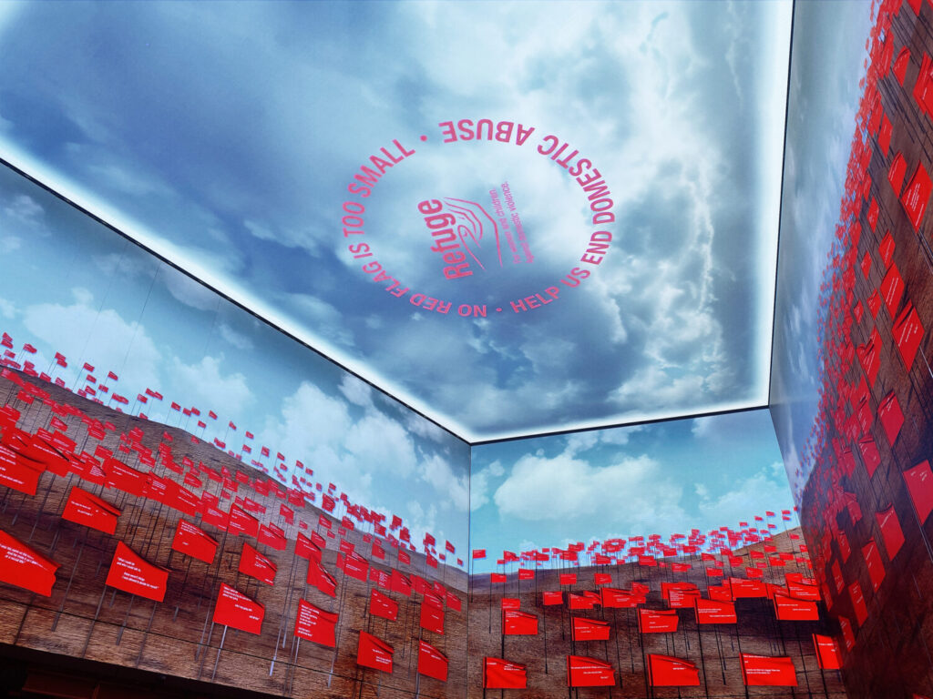 Photo of the Outernet, London. Looking towards the ceiling, three walls are filled with images of red flags, and on the ceiling is the sky with Refuge's logo encircled by the text 'No red flag is too small. Help us end domestic abuse.'